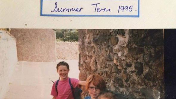 A young Josie Russell stood next to a stone wall with two other pupils, and smiling at the camera. She is wearing a pink t-shirt and has a blue rucksack on her shoulders. Her brown hair is tied back.