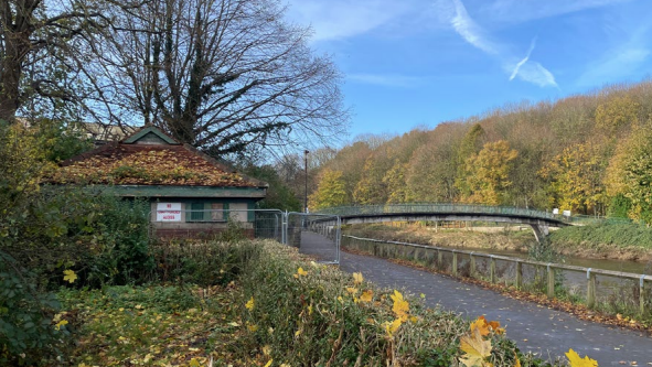 The old toilet block is a one story brown-brick building with a triangular roof, green door and  window panes. There is a pile of yellow leaves on the roof. NO UNAUTHORISED ACCESS is written in red on a sign attached to the fenced-off building. There is a promenade next to it with the arched Baths Bridge visible in the distance. There are many trees across the river, with their leaves falling off. 