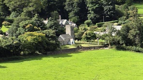 Oare Church near Malmsmead. The building is surrounded by trees in full bloom and green fields. Other buildings are partially hidden by the trees in the area.