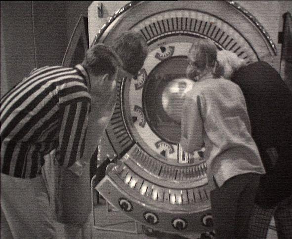 Screenshot of the backs of Ian, Barbara, Vicki and The Doctor all watching an image appear on a screen at the centre of a contraption with many switches and dials