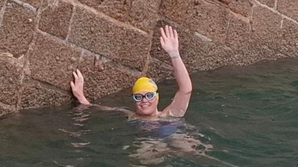 Marie-Helene Sanderson in the water. She is wearing goggles and a yellow swim hat and is waving at the camera while smiling broadly. Her other hand is on a stone sea wall.