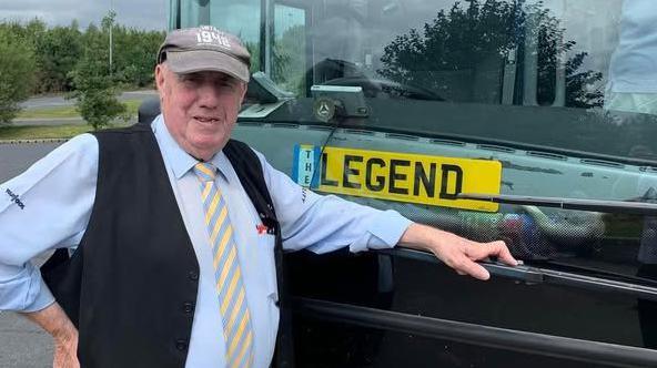 A man in a cap, waistcoat, shirt and tie standing in front of a coach. There is a number plate in the window with "legend" printed on it.