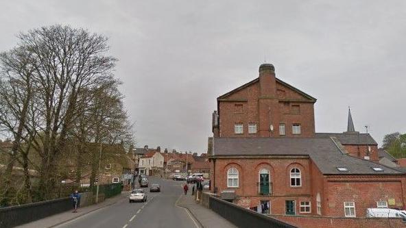 A road bridge with a large redbrick building on the right-hand side of the image, with a pavement and railings flanking the road. Cars and pedestrians are in the far distance.