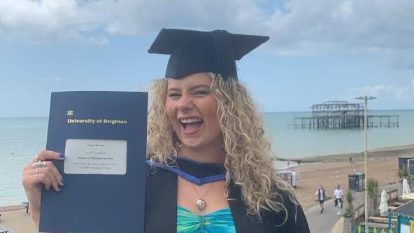 A girl with blonde curly hair winks at the camera holding her graduation certificate and wearing black robes and a hat