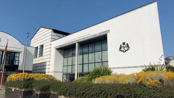 Douglas Courthouse building, which is a large white glass-fronted building with the government crest on the right and the words Isle of Man Courts of Justice in black lettering above the entrance. Planters containing yellow-flowering gorse are sitting in front on the building.