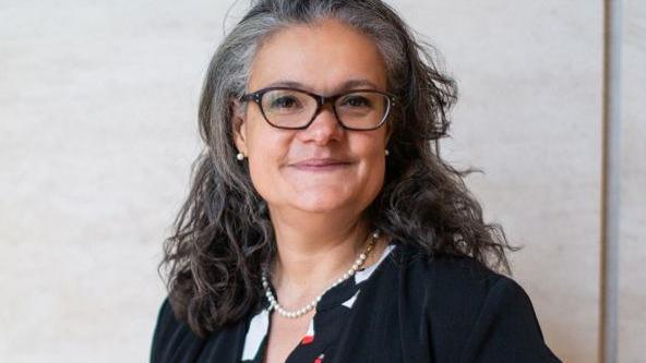 Emily Holzhausen has wavy brown and greying hair. She is wearing pearl earrings and necklace, glasses and a black jacket and white, red and black patterned top.