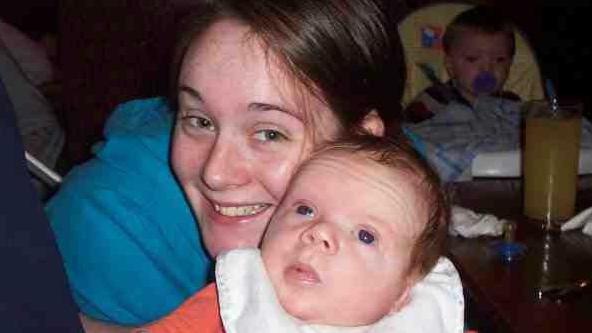 Becci Smart holding Zak when he was a baby - she is behind him in a blue top and has her dark hair pulled back in a ponytail and has a big smile on her face. Zak is a young baby with an orange top on and a white bib over the top, he has very short fluffy baby hair and is looking up at the roof. There is a baby in a high chair with a dummy in his mouth seen in the background of the image.