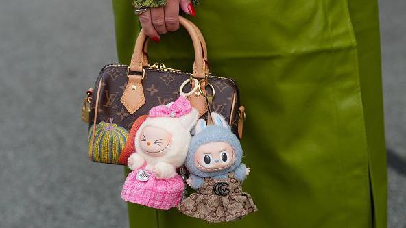 The dolls have become a style accessory and people are decorating their bags with them as charms. A woman carries a Louis Vuitton handbag with two dolls fastened to the handle. the dolls have grey and cream fur and have been given brown and pink dresses. One of the dolls has stitching for eyes and a pink bow on its head.