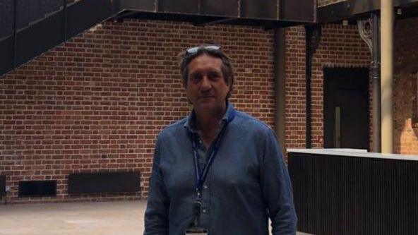 Matt Smith stood in the new bar area with exposed brick on the walls and a large black staircase behind him. He is wearing a blue shirt with a blue lanyard and sunglasses on his head.