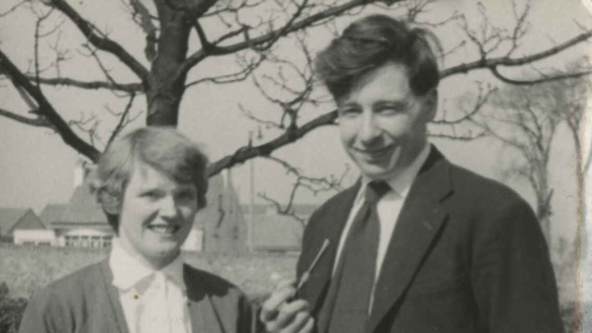 Jean Purdy and Prof Edwards pose outdoors in a vintage setting, framed by a leafless tree and distant buildings. Ms Purdy wears a cardigan and skirt, while Prof Edwards is dressed in a suit and tie, holding a pair of glasses. 