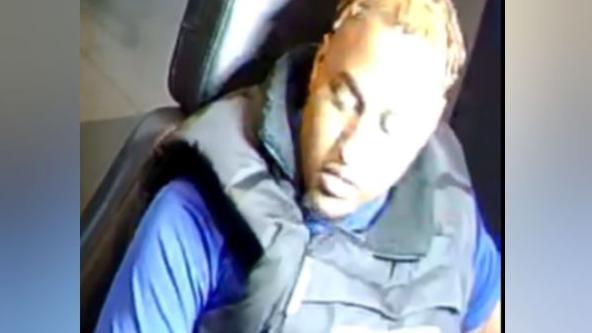 A CCTV image of a man with blond hair wearing a black quilted body warmer and blue short-sleeved T-shirt, sitting down in a black or dark-coloured leather-looking chair