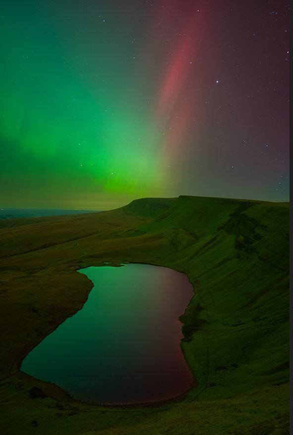 Llyn y fan Fach in the Bannau Brechaunog national park in Carmarthenshire is surrounded by a steep ridge on the right hand side and mountains beyond. The lake is bathed in green and red light reflected from the skies above.