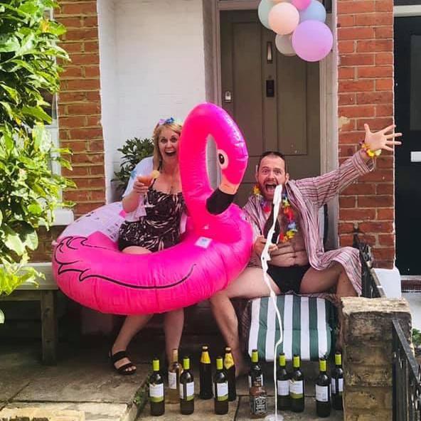 Beth, in sunglasses, perches on a large pink flamingo inflatable beside Tom, who wears a robe and swim shorts. They sit outside their front door surrounded by wine bottles, with pastel-coloured balloons strung overhead.
