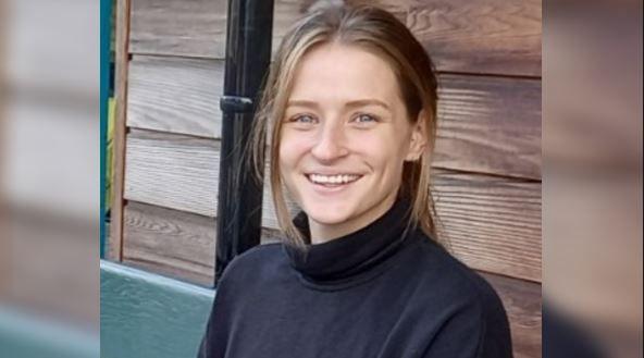 A young woman with light brown hair in a ponytail, wearing a black turtle neck and blue jeans sits on a bench. She is smiling at the camera.
