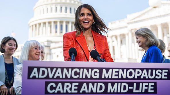 Halle Berry seen in red suit with the US Capitol in the background. She has a mic in front of her and board that says Advancing Menopause Care and Mid-Life Women's Health Acts