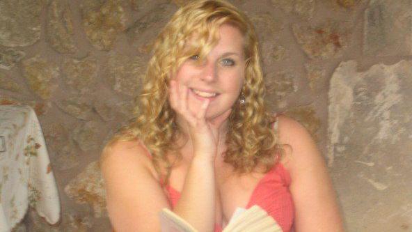 Melissa Power sits holding an open book and and looks at the camera smiling. She has long blonde curly hair and is wearing a coral-coloured skimpy top. She is inside a building with rough stone walls and the corner of a table with a table cloth is to the left of the picture.
