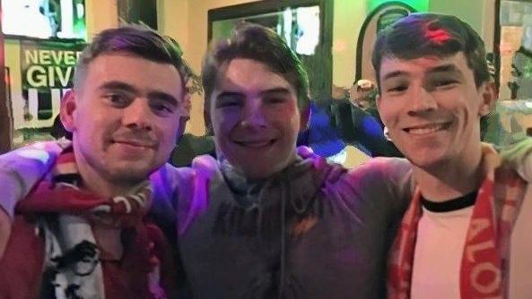 Connor, Lucas and Bradley, three young men smile, they all have dark hair, and Connor and Bradley wear football scarves as they stand in a pub.
