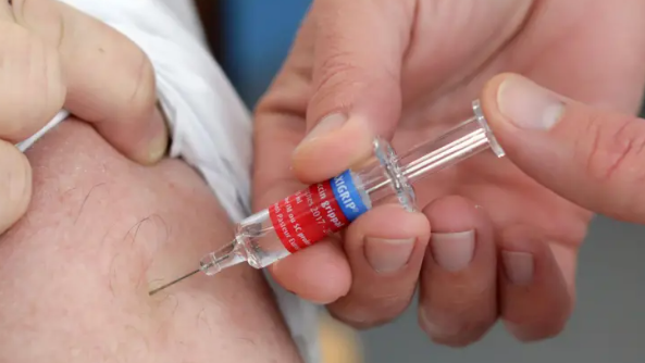 A man receiving a vaccination into his arm. He is wearing a white t-shirt while the medic is pressing down on the syringe to administer the vaccine.