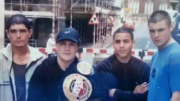 A dated photograph of Ricky Hatton with a championship belt draped over his shoulder posing with three other male boxers on a street. They are standing in casual clothing in front of a building site that is covered in scaffolding.