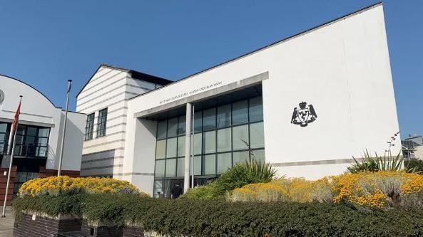 The Isle of Man Courts of Justice, which is a large white glass-fronted building with the government crest on the right and the words Isle of Man Courts of Justice above the entrance.