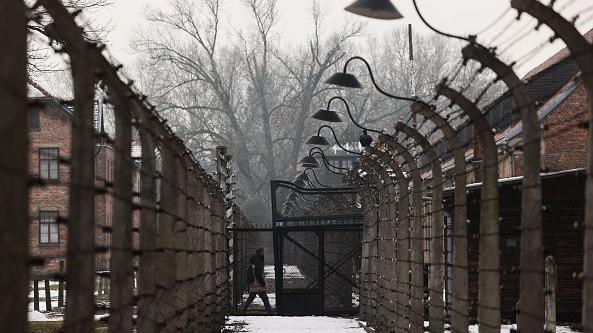 A visitor at the former Nazi German concentration camp Auschwitz-Brikenau. There are large, dark gates and towering fences, and buildings on either side of the barriers, with wintry trees ahead. There is snow on the ground.