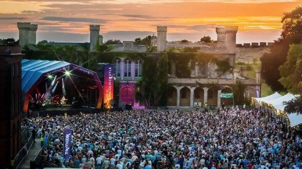 An outdoor concert in front of an ivy-covered castle with towers. A brightly lit stage on the left has a musician performing, while a large crowd are stood in front. The sky glows with warm sunset colors of yellows and pinks.