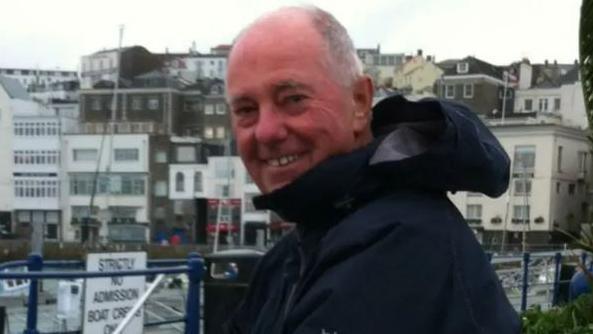 A head-and-shoulders image of Jack Honeybill. He is smiling directly at the camera and is wearing a navy raincoat. The harbour is shown in the background and it is a cloudy day.