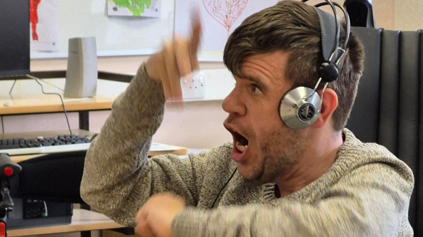 A white man with brown hair and stubble wearing headphones looking at a computer with a computer in the background. It is a side angle and a head and shoulders shot of him. 
