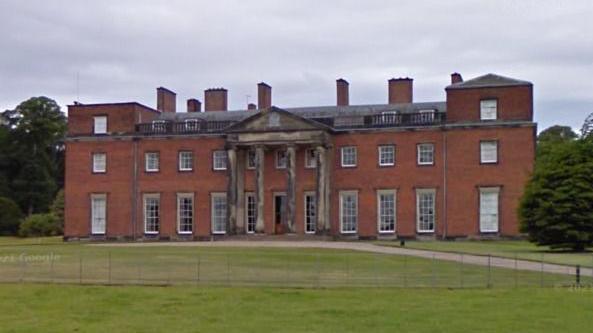 A street view image of a large 18th Century country hall. It is made up of brown bricks and has a large set of columns near its entryway that have seen better days.