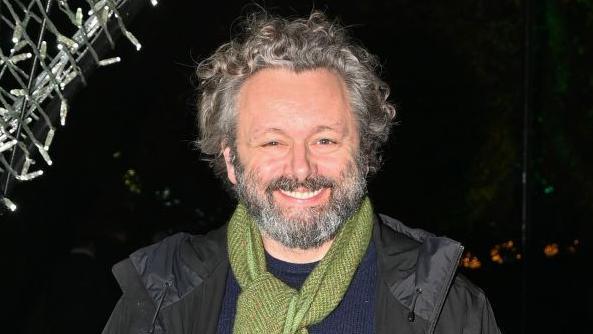 A head and shoulders shot of Michael Sheen. He is stood outside and it is dark. He has short grey, curly hair and a grey beard and moustache. He has a black jacket, blue jumper and green scarf on.