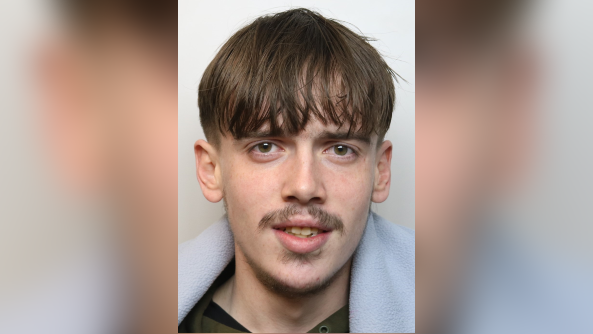 Leo Morris looking at the camera in a police mugshot. He has a long fringe that looks greasy and a straggly moustache. He has yellow teeth.