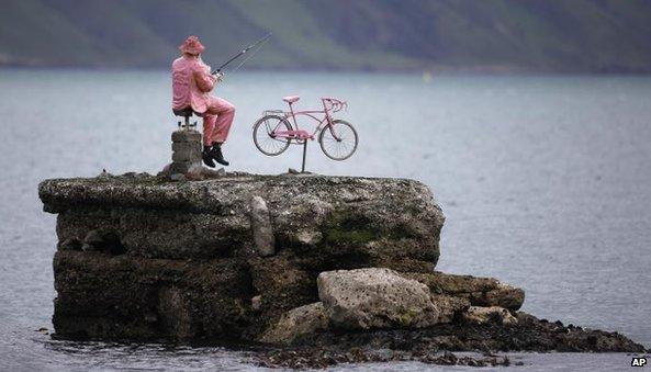 A pink statue of a man fishing next to his bike sits on the rocks at Waterfoot