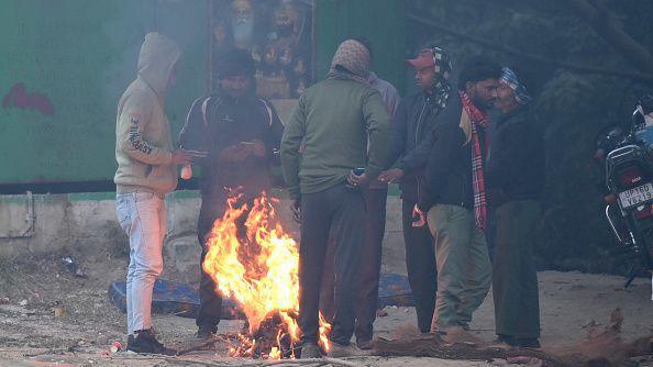 Seven men are standing around bonfires, wearing sweaters and jackets to protect themselves from the cold.