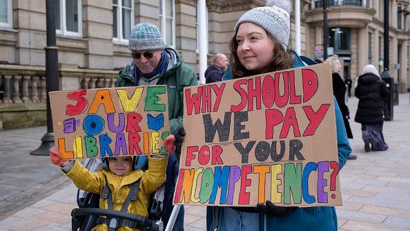 A man, woman and child in a pram stand with colourful home-made signs at a protest against council cuts. The signs read "Save our Libraries" and "Why should we pay for your incompetence?"