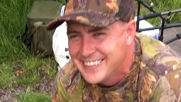 Paul Smith wearing a camouflage patterned t-shirt and cap with grass and a olive green bag in the background. He is smiling.