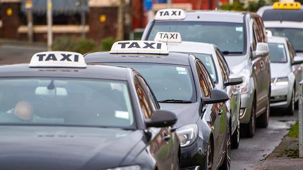 A row of taxi vehicles parked up at the side of the road. They all have a white sign on the roof of their car, apart from one at the back which has a yellow sign.