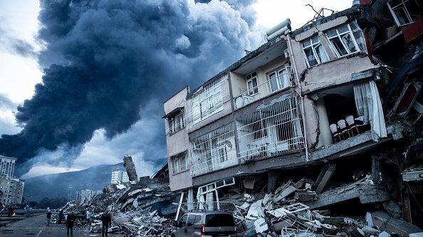 moke billows from Iskenderun Port fire as people walk past collapsed buildings in Iskenderun, Turkey.