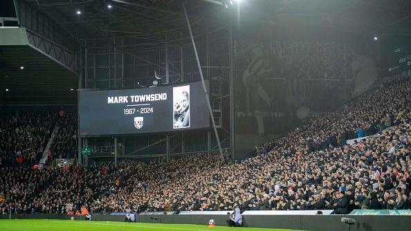 A large screen with the words Mark Townsend and a black and white picture at a full football stadium