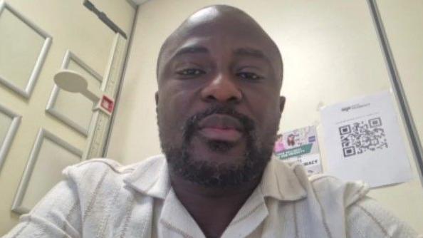 A close-up of a black man staring straight ahead. He has very short black hair and a short beard and is wearing a white shirt.