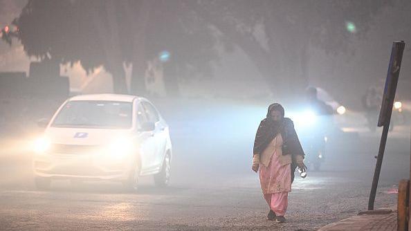 A woman wearing a pink traditional Indian dress and a black scarf walks along the side of the road in the early hours of the morning in Karol Bagh, Delhi, as vehicles pass by with their lights on due to low visibility.