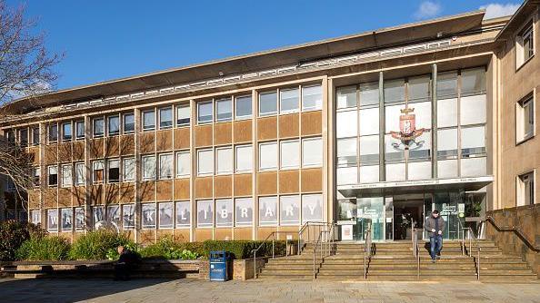 A large flat roofed building with a mostly glass facade. There is an entrance with stairs leading to it on the right side, above the entrance is a coat of arms