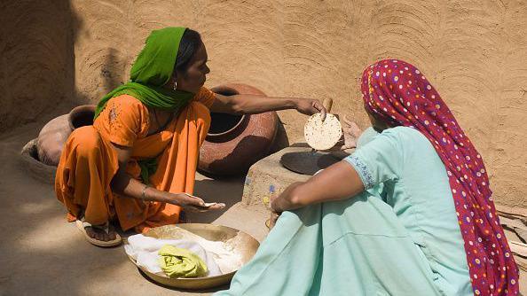 Two women seen sitting in the sun as they make Indian flatbread on a stove. One of them is dressed in orange salwar-kameez with a green dupatta on her head. The other wears a pale blue salwar-kameez with a maroon dupatta with colourful dots.  