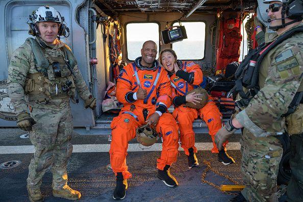 The two Nasa astronauts Victor Glover and Christian Koch lean against each other in orange jumpsuits with big smiles flanked by camouflaged military personel. Victor gives a thumbs up.