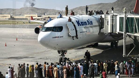 Dozens of people queue on the tarmac, while others stand or sit on the roof of a passenger plane