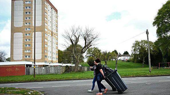 Local residents take wheeled bins of uncollected rubbish to dustbin and refuse lorries at a temporary collection point set up in Shard End on 14 April. Two people are walking together with their bins and talking as they go.
