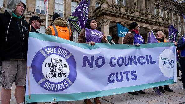 Protesters wearing Unison-branded tabards and jackets carry a large blue and green banner at a protest outside Birmingham Council House.