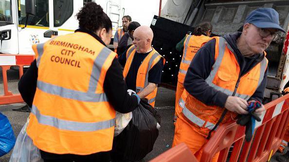Residents hand rubbish to refuse workers at a Mobile Household Waste Centre in Senneleys Park on 8 April. Refuse workers are wearing orange high-vis jackets with Birmingham City Council written on the back. Barriers are in place to keep the area clear.