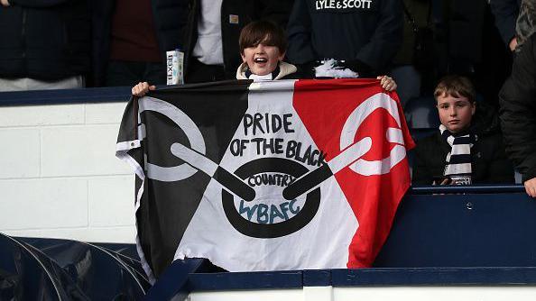 A young boy with short brown hair, standing in a football stand at West Bromwich Albion and holding a Black Country flag. The flag has black, white and red sections and and black looped chain across the middle. The boy is smiling towards the camera. 