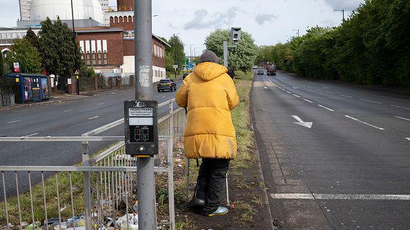 Homeless man on crutches stands beside a main road waiting for cars to pull up at the lights.