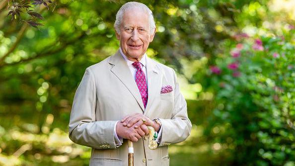 King Charles III is photographed in the Arboretum at Highgrove House with trees and bushes dappled in sunlight blurred out in the background behind him. He wears a pale suit jacket with a pink pocket square and pink tie. His hands are resting on top of a cane and he is smiling.